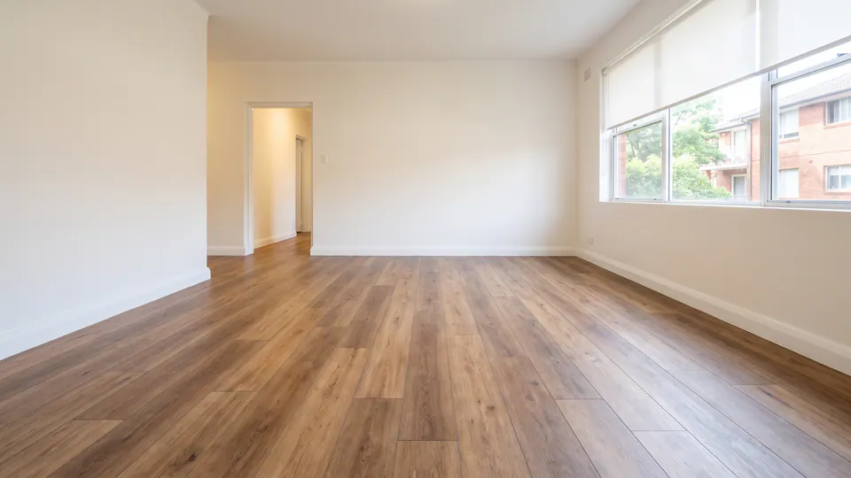 An empty apartment living room with a timber-look plank floor, lit by soft morning daylight from a side window