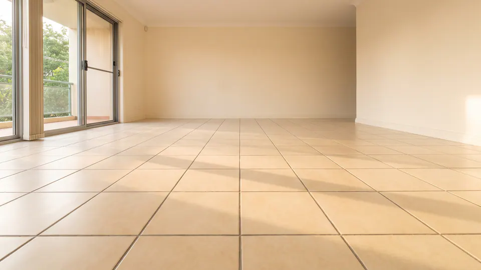 An empty living area with an existing cream tiled floor, its grout lines lit by low daylight from a sliding door