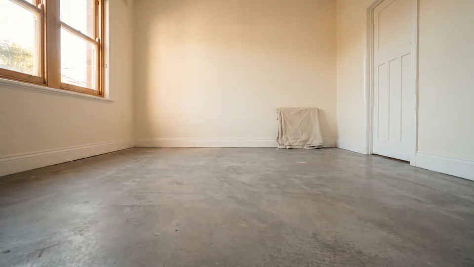 An empty living room with its old floor covering removed, showing the bare concrete slab lit by daylight from a side window